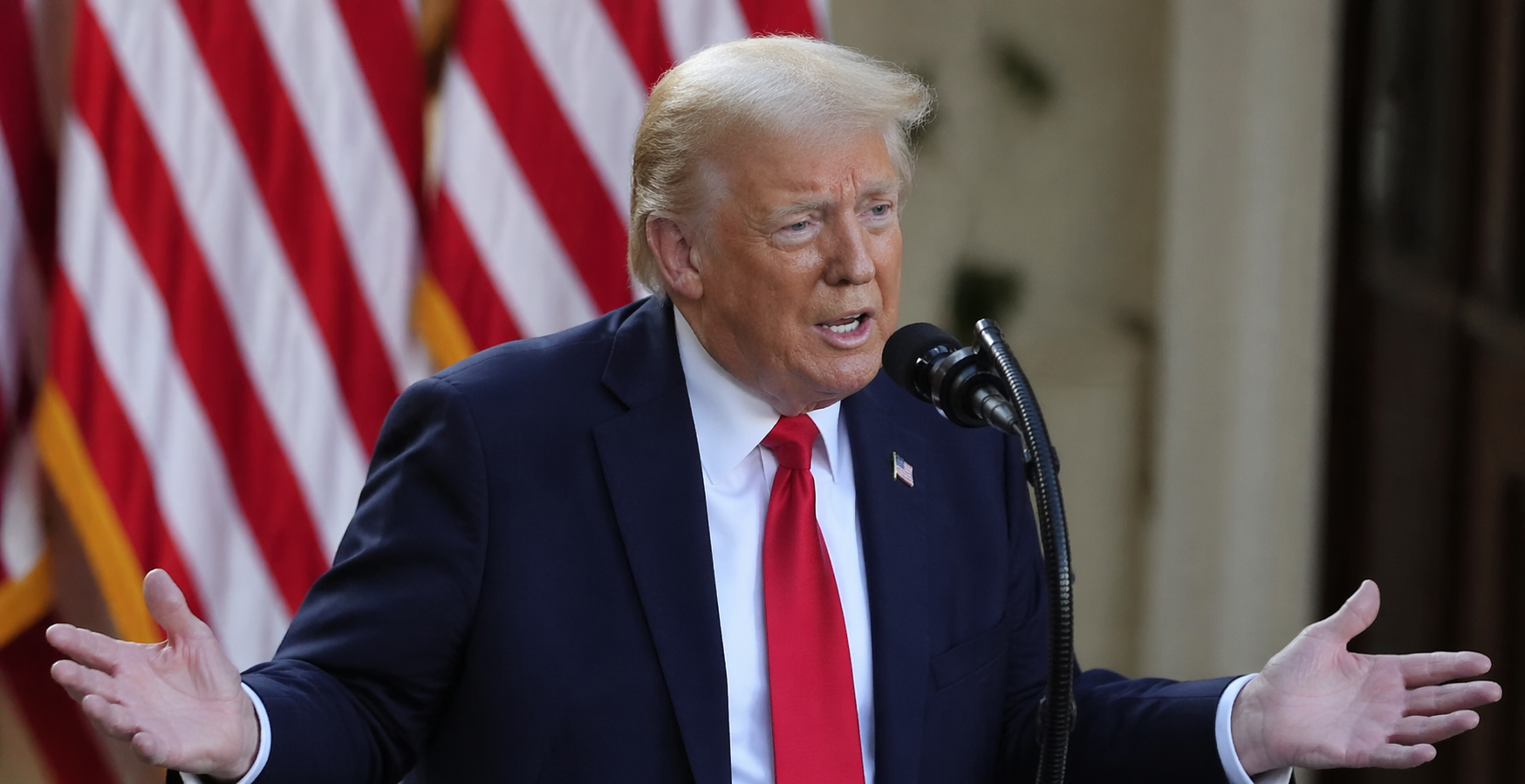 Donald Trump speaks at a microphone in front of US flags, gesturing during a formal public address.