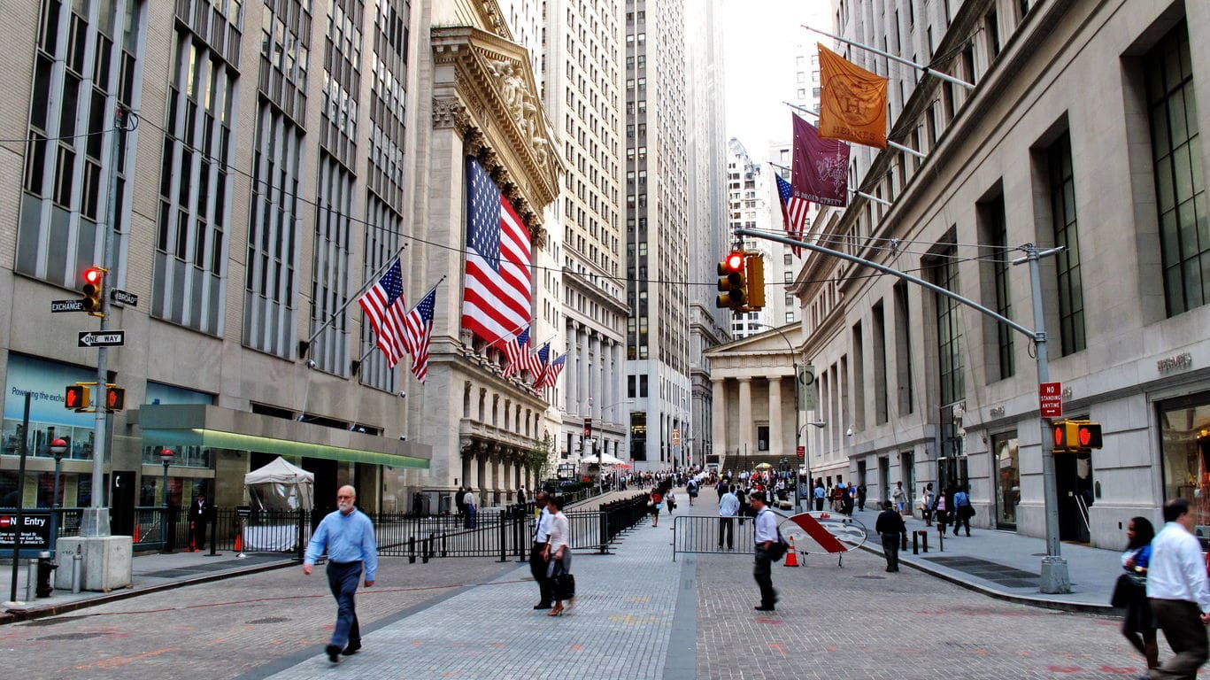 Pedestrians walk through Wall Street in Lower Manhattan beneath American flags near the New York Stock Exchange.