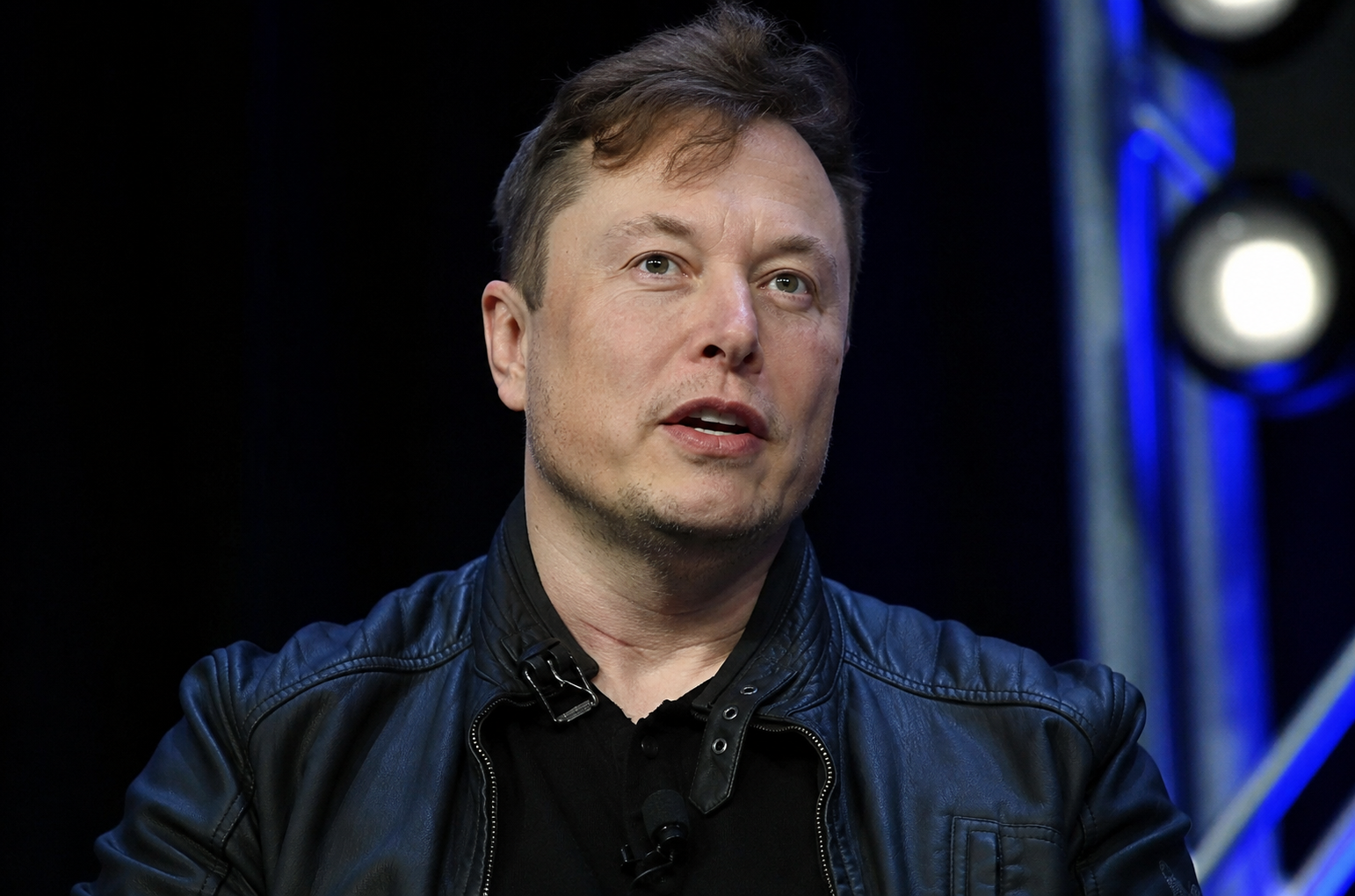Elon Musk speaks on stage while wearing a black leather jacket.