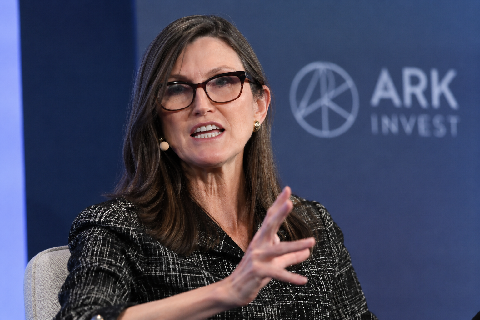 Cathie Wood speaks during an ARK Invest event, gesturing on stage with the firm’s logo visible behind her.