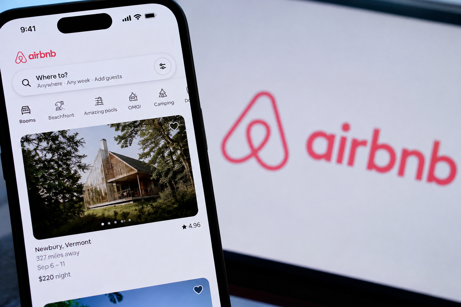 Airbnb app displayed on a smartphone with the company logo visible on a screen behind it.