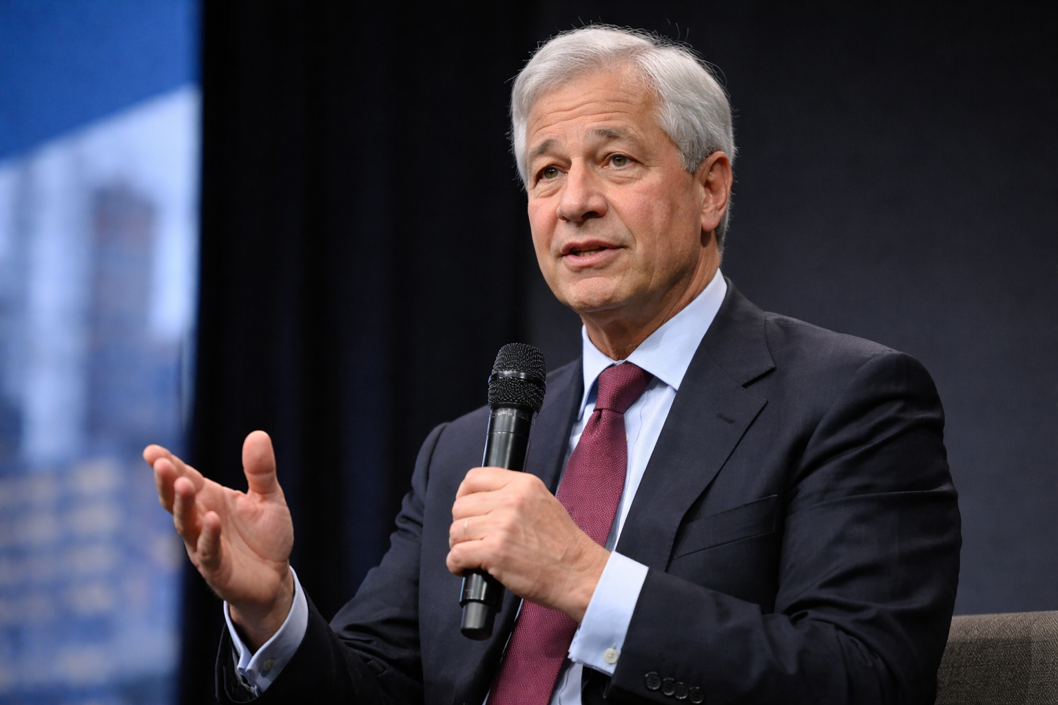 Jamie Dimon speaks on stage at an investor event, gesturing during a discussion on markets, banking and the economy.