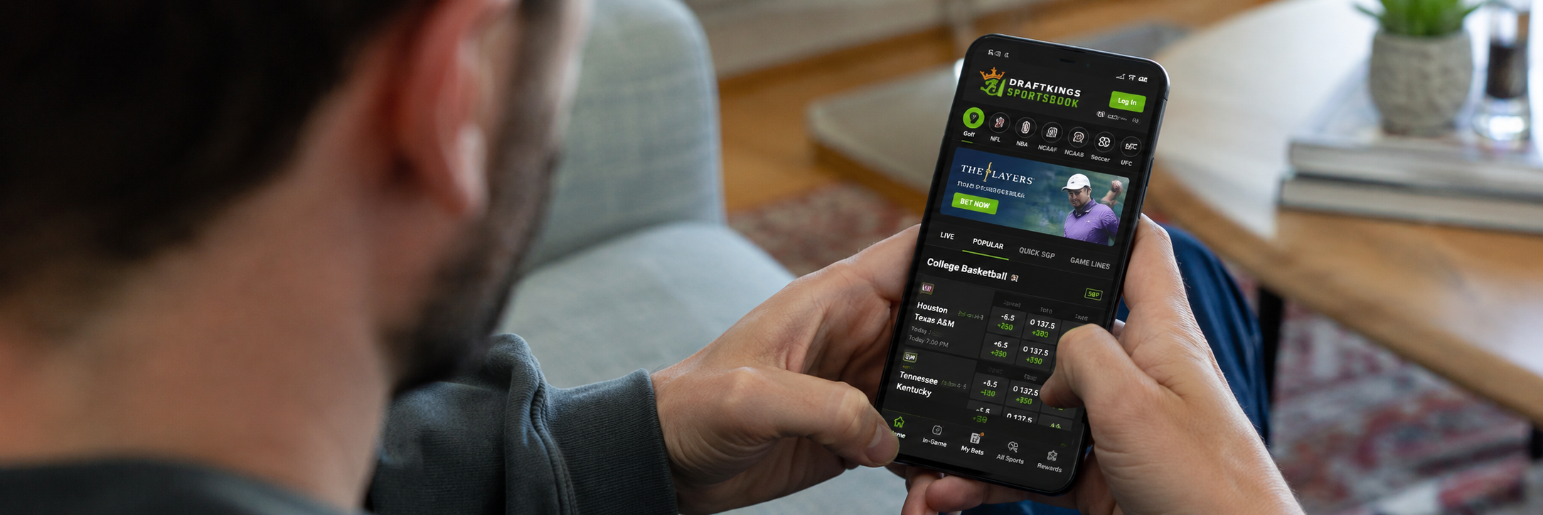 An over-the-shoulder view of a person holding a smartphone displaying the DraftKings Sportsbook app with live betting lines for college basketball.