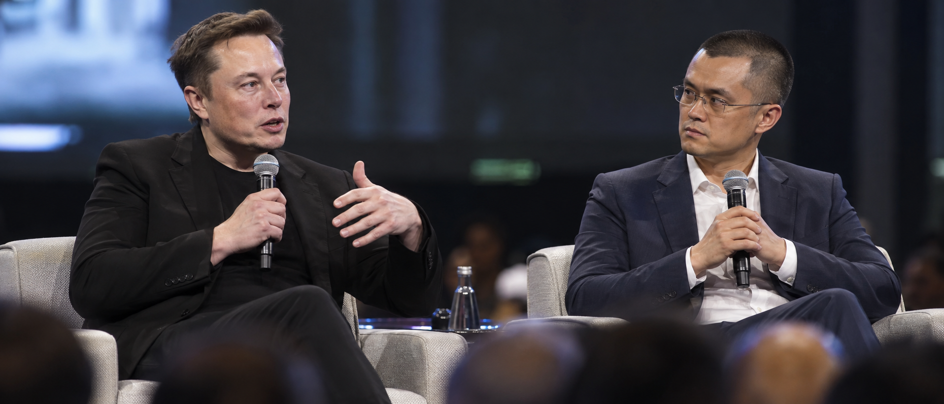 Elon Musk and Changpeng Zhao seated on armchairs on a stage holding microphones, with Musk speaking and gesturing while Zhao listens.