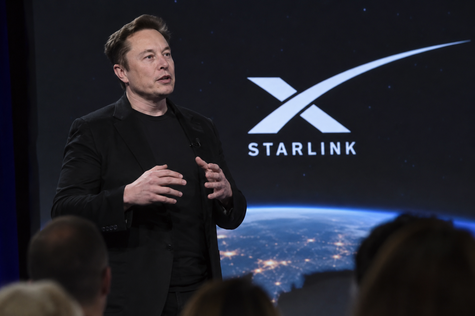 Elon Musk stands on stage in front of an audience, gesturing while delivering a presentation with the SpaceX Starlink logo and a view of Earth displayed on a large screen behind him.